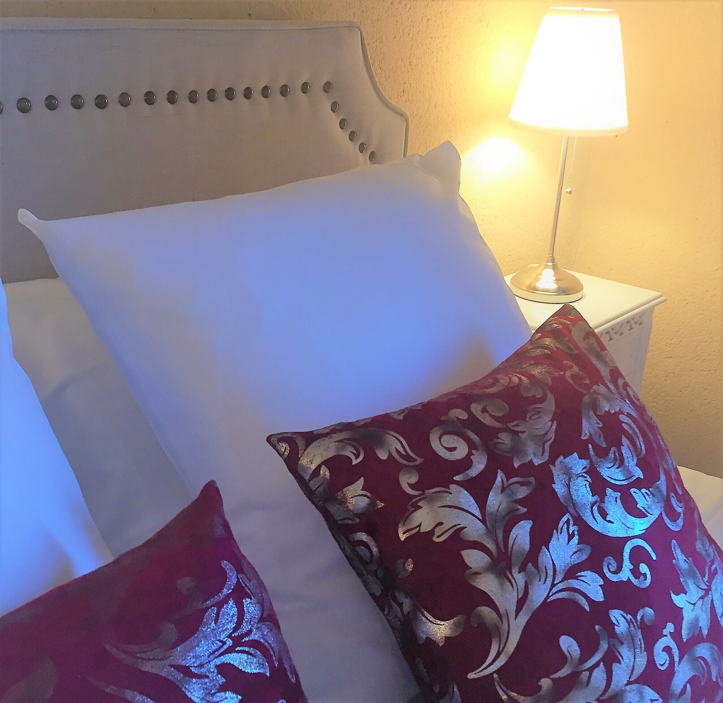 A neatly made bed with white and red pillows, a white headboard, and a bedside lamp.