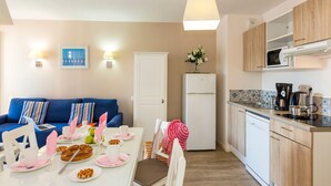 A modern kitchen with a white refrigerator, a microwave, and a coffee maker. There is a dining table set with plates, glasses, and a cake. A blue sofa and a white chair are visible in the background. - Studio cosy et équipé, près de la plage (Pornichet)