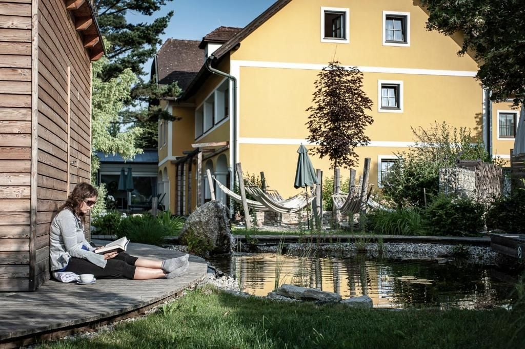 A person sitting on a wooden deck reading a book, with a pond and a yellow building in the background.