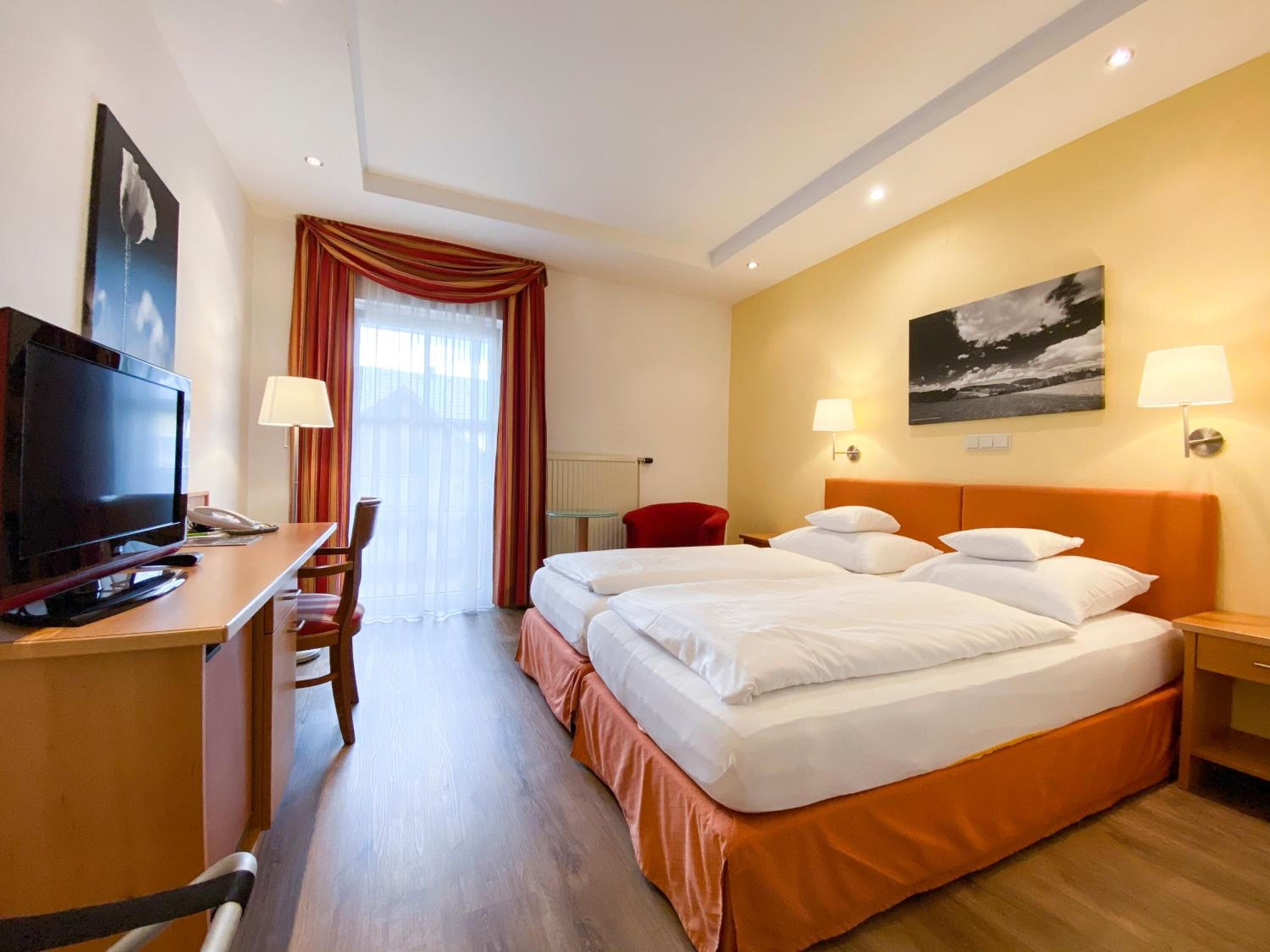 Una habitación de hotel con una cama, un escritorio con televisor, una silla, una lámpara y un cuadro en la pared.