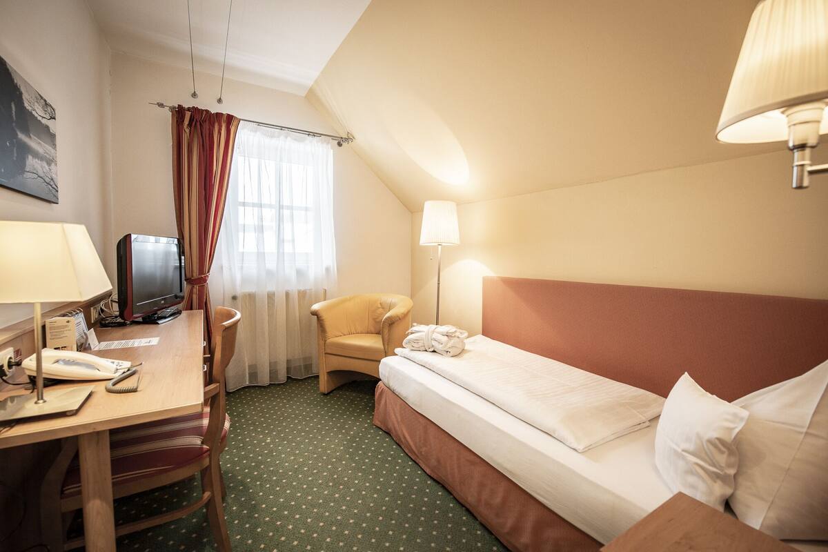 Una habitación de hotel con cama, escritorio con computadora, silla, lámpara y ventana con cortinas.