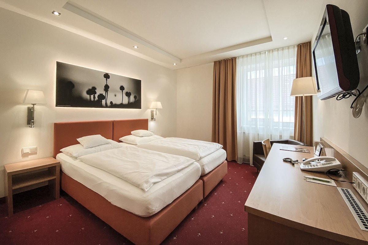 Una habitación de hotel con dos camas, un escritorio con teléfono y lámpara, un televisor montado en la pared y un cuadro sobre las camas.