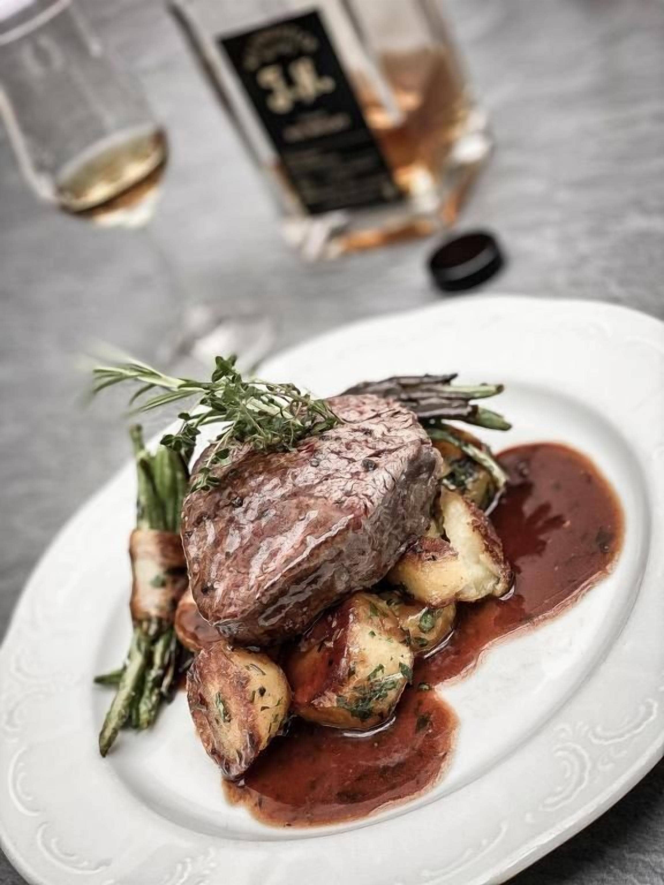 A plate with a steak, potatoes, and a sauce, accompanied by a glass of wine and a bottle of liquor.