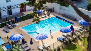 Piscina exterior con sillas de descanso y sombrillas, rodeada de una valla y árboles.
