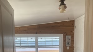 A bedroom with a large window overlooking a beach, wooden walls, a bed with a quilt, and a ceiling light fixture. - River Meadow Camp Cottage (Summerside)