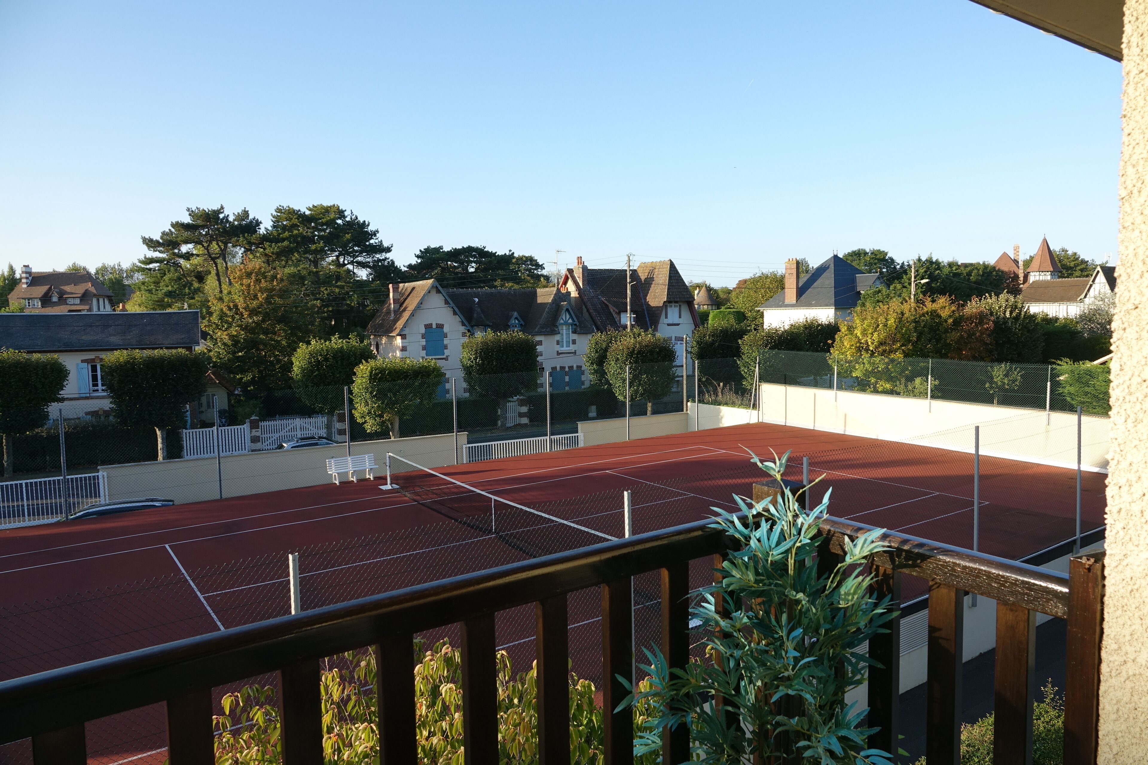 Un campo da tennis con recinzione in legno, circondato da case residenziali e vegetazione.
