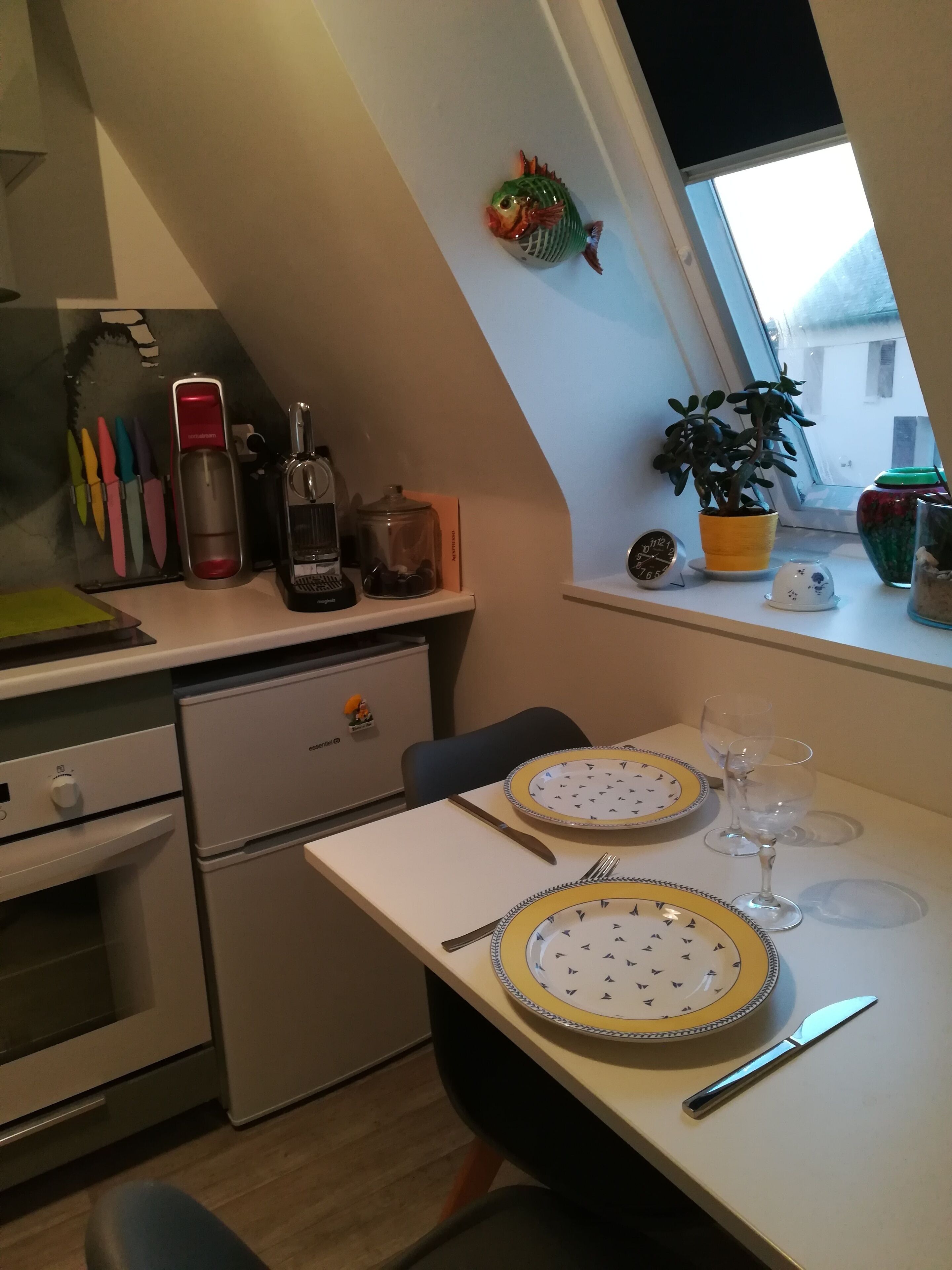 A compact kitchen with a dining area, featuring a small refrigerator, a coffee machine, and a window with a view.