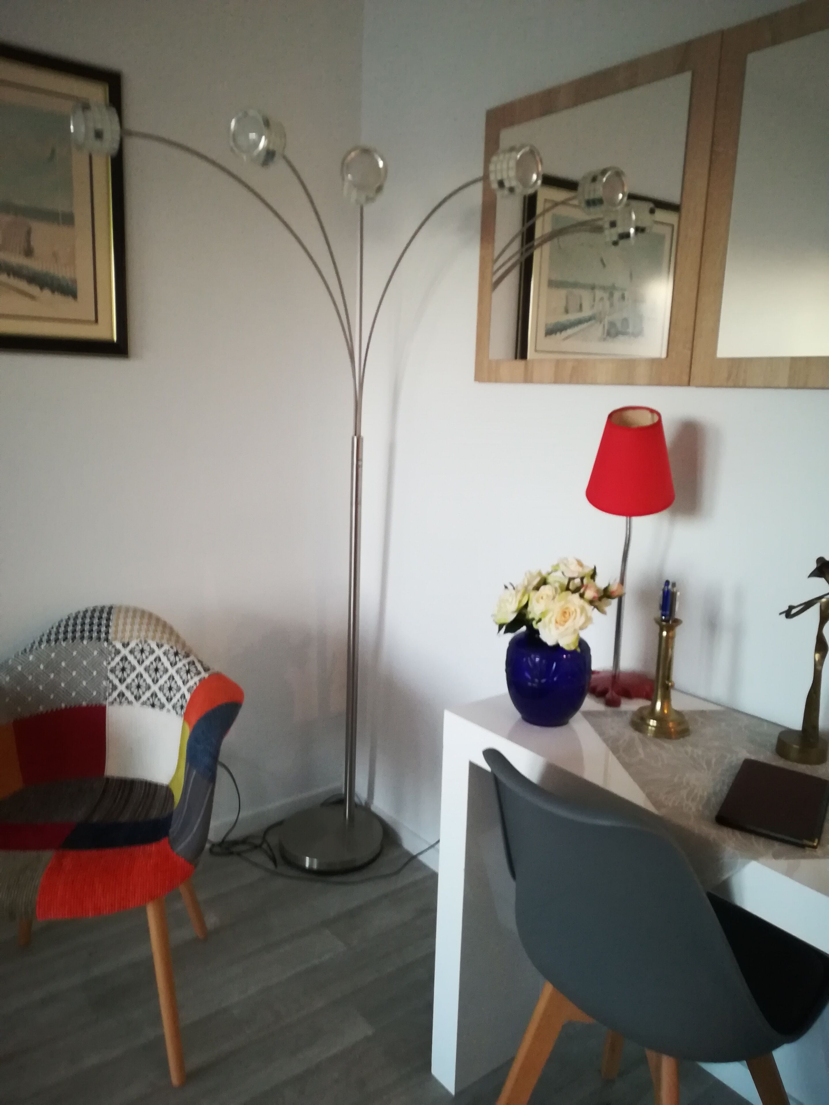 Une chambre moderne avec un bureau blanc, une lampe rouge, un vase avec des fleurs, une chaise avec un coussin coloré et un grand lampadaire.