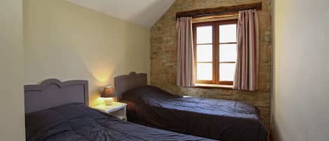 A bedroom with two beds, a nightstand, a lamp, a window with curtains, and a stone wall.