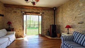 A rustic room with a stone wall, wooden floor, a sofa, a striped couch, a fireplace, and a large window with a view of greenery.