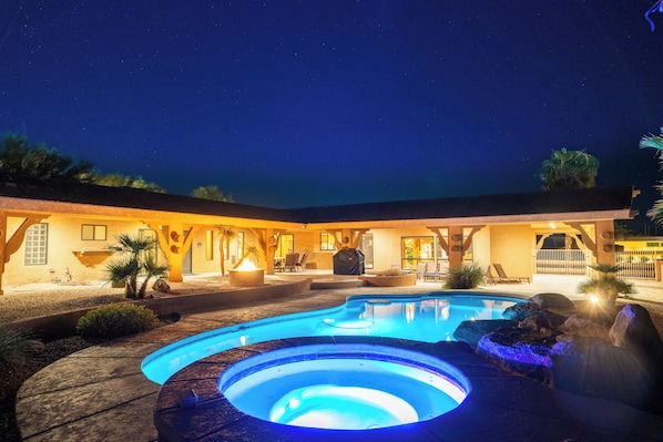 A nighttime view of a pool area with a hot tub, surrounded by a building with outdoor seating and a patio.