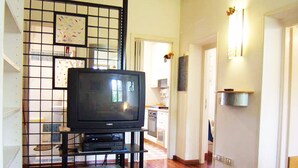 Sala de estar com televisão em um móvel, cozinha ao fundo e corredor que dá para outro quarto.