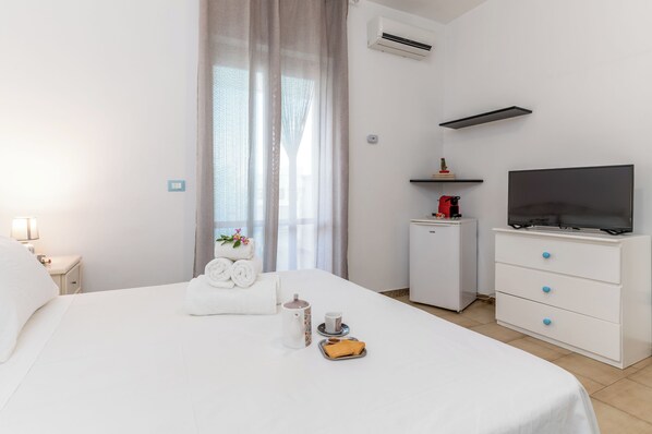 Habitación de hotel con cama, televisión, refrigerador pequeño, escritorio y ventana con cortinas.
