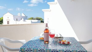 Una mesa con una botella y vasos, un cuenco de aceitunas y un plato de fruta en un balcón, con un edificio blanco al fondo.
