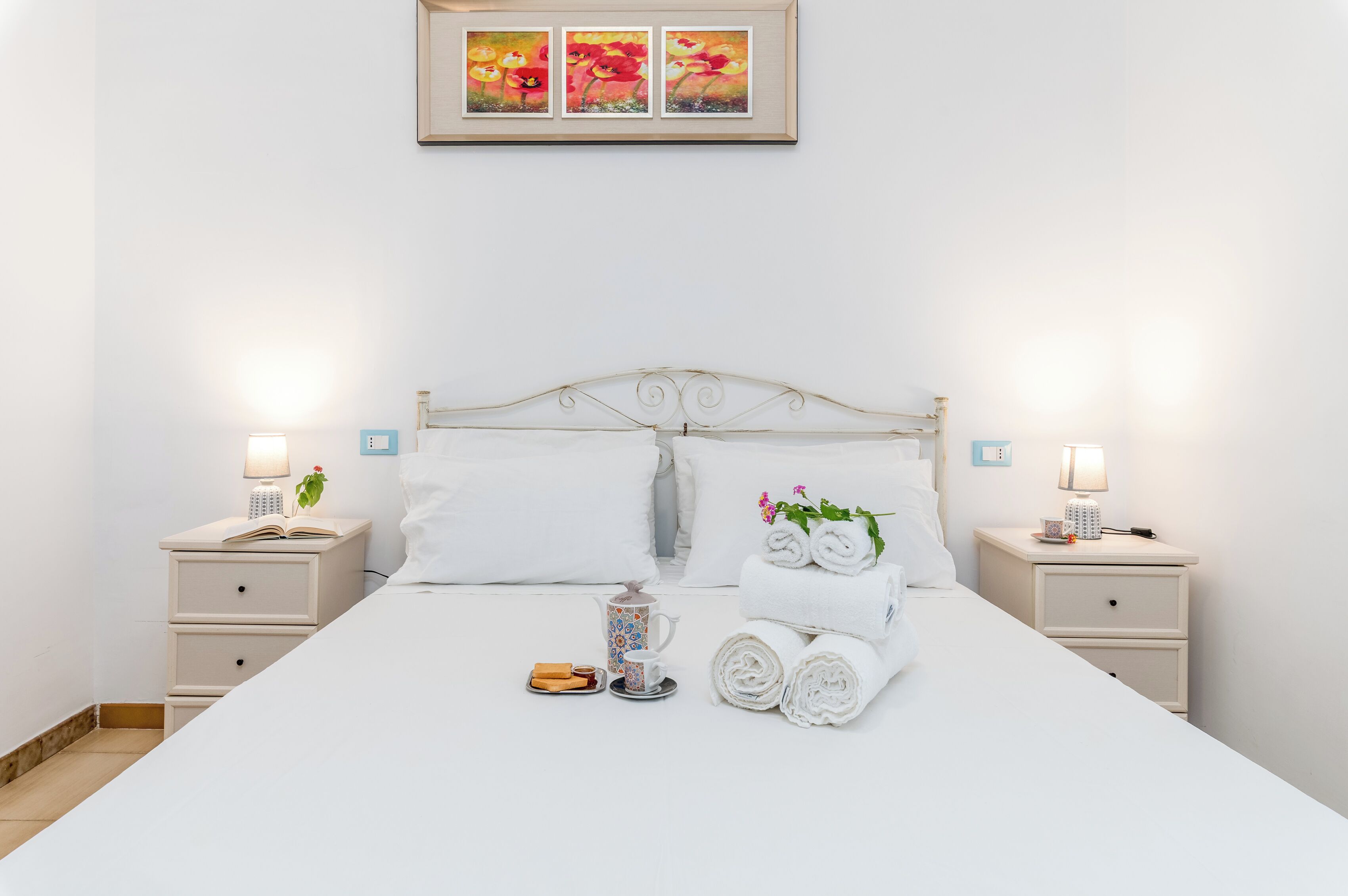 Een opgemaakt bed met witte lakens, twee nachtkastjes met lampen, een ingelijst beeld boven het bed, en een klein tafeltje met toiletartikelen.