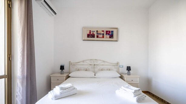 Habitación de hotel con una cama, dos mesitas de noche, aire acondicionado de pared, una ventana con cortinas y un cuadro enmarcado sobre la cama.