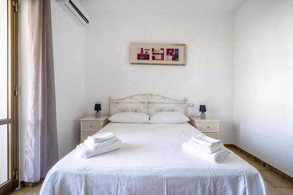 Habitación de hotel con una cama, dos mesitas de noche, aire acondicionado de pared, una ventana con cortinas y un cuadro enmarcado sobre la cama.