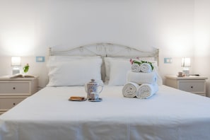 Un letto rifatto con lenzuola bianche, una teiera decorativa e asciugamani arrotolati su una testiera bianca.