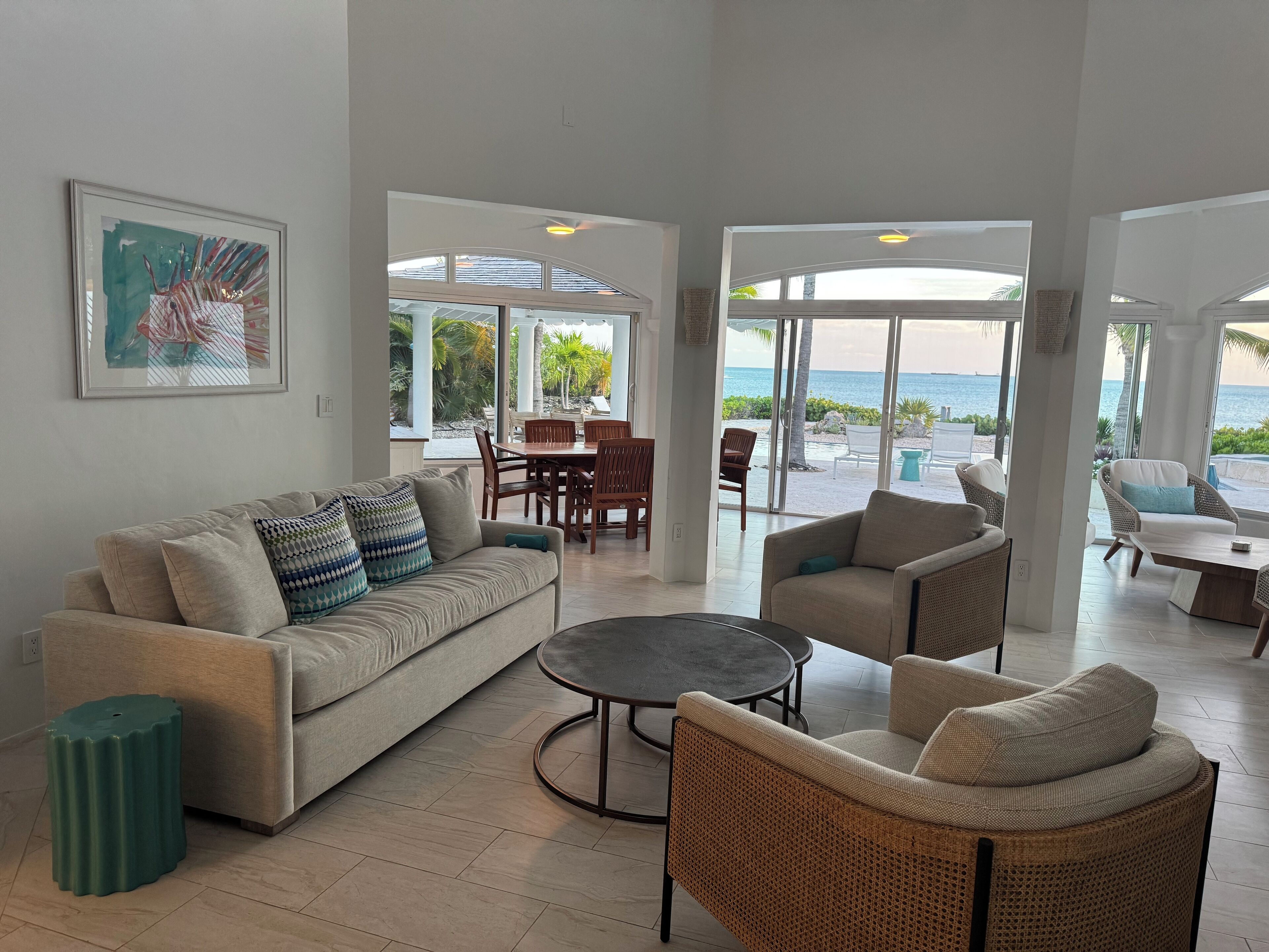 A spacious living area with a sofa, chairs, and a coffee table. There is a dining table with chairs and a view of the ocean through large windows.