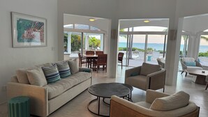A spacious living area with a sofa, chairs, and a coffee table. There is a dining table with chairs and a view of the ocean through large windows.
