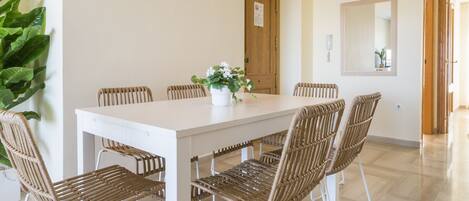 Un comedor con una mesa blanca y sillas de mimbre, una planta en maceta sobre la mesa, y una puerta de madera al fondo.