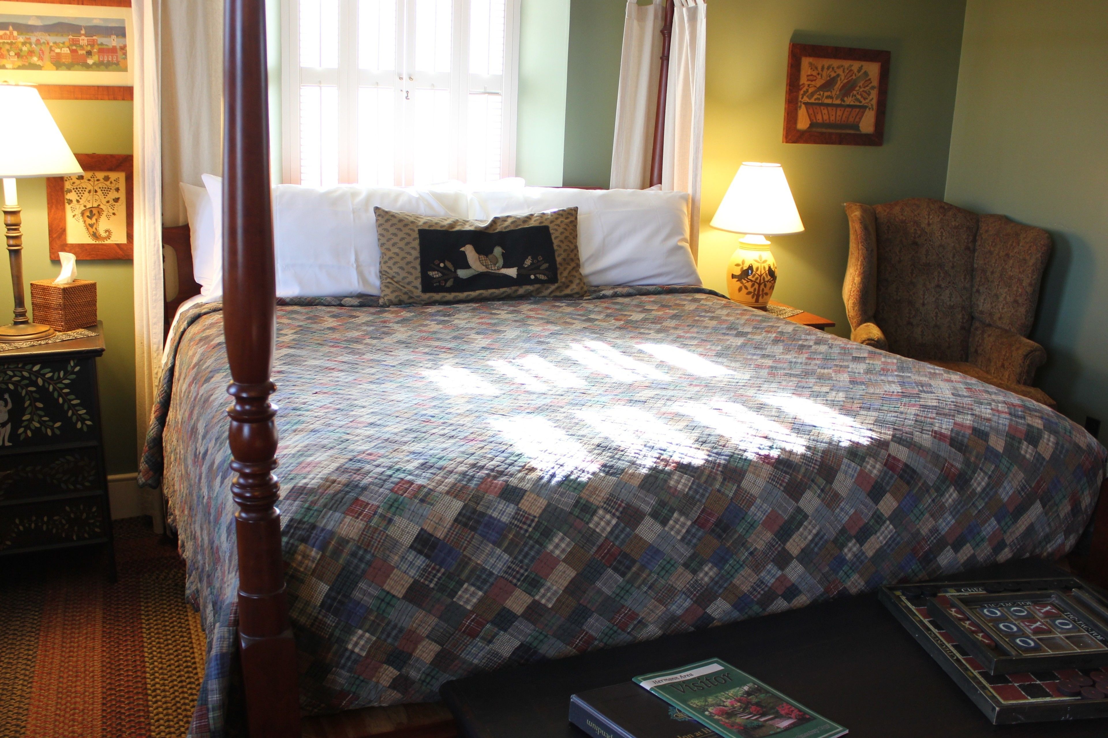 A neatly made bed with a plaid bedspread, a wooden headboard, and a single decorative pillow. There is a chair and a small table with a lamp and a book beside the bed.