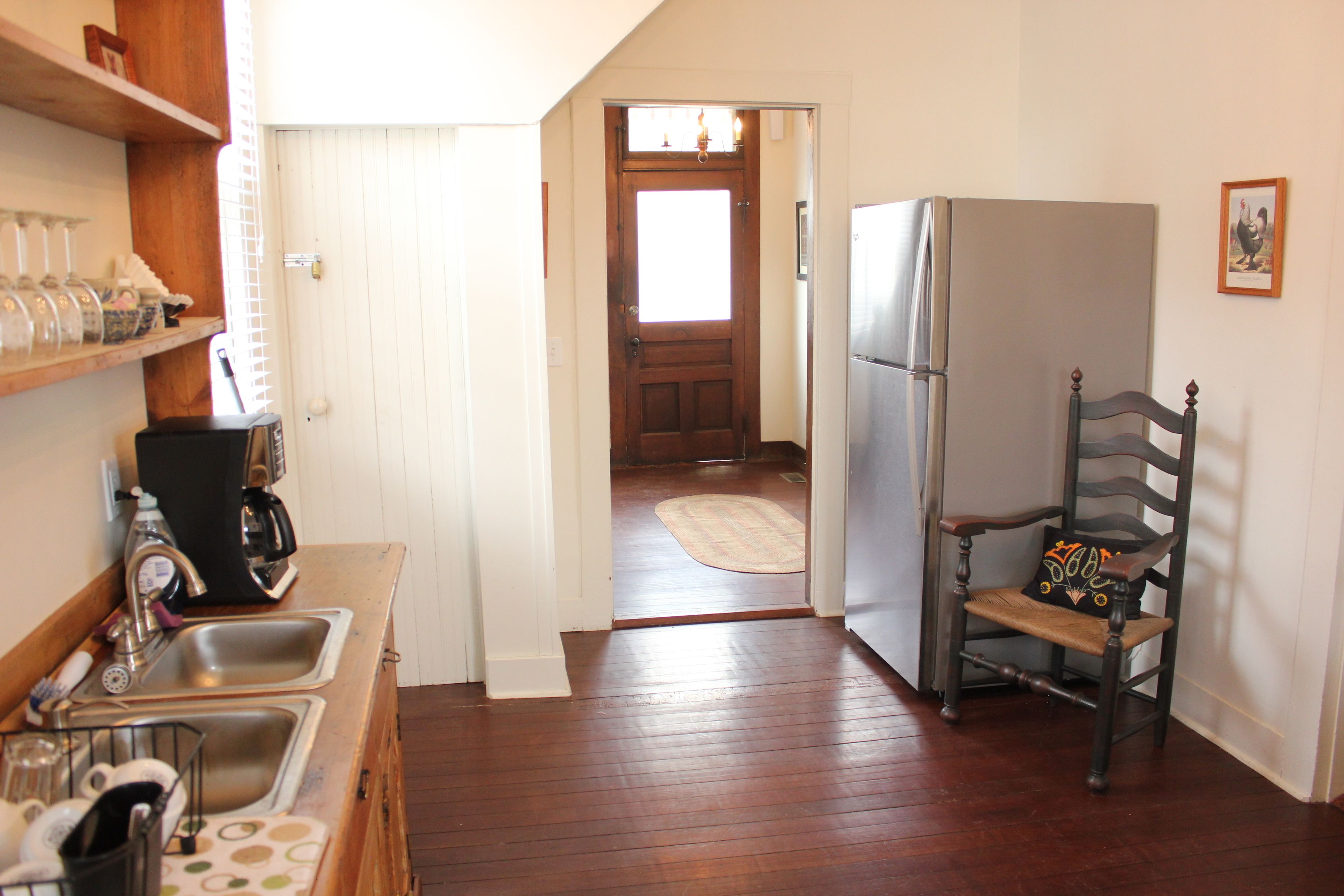 A kitchen with wooden floors, a refrigerator, a chair with a decorative pillow, and a door leading to another room.