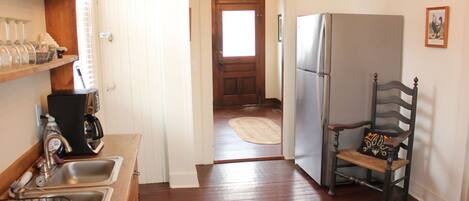 A kitchen with wooden floors, a refrigerator, a chair with a decorative pillow, and a door leading to another room.