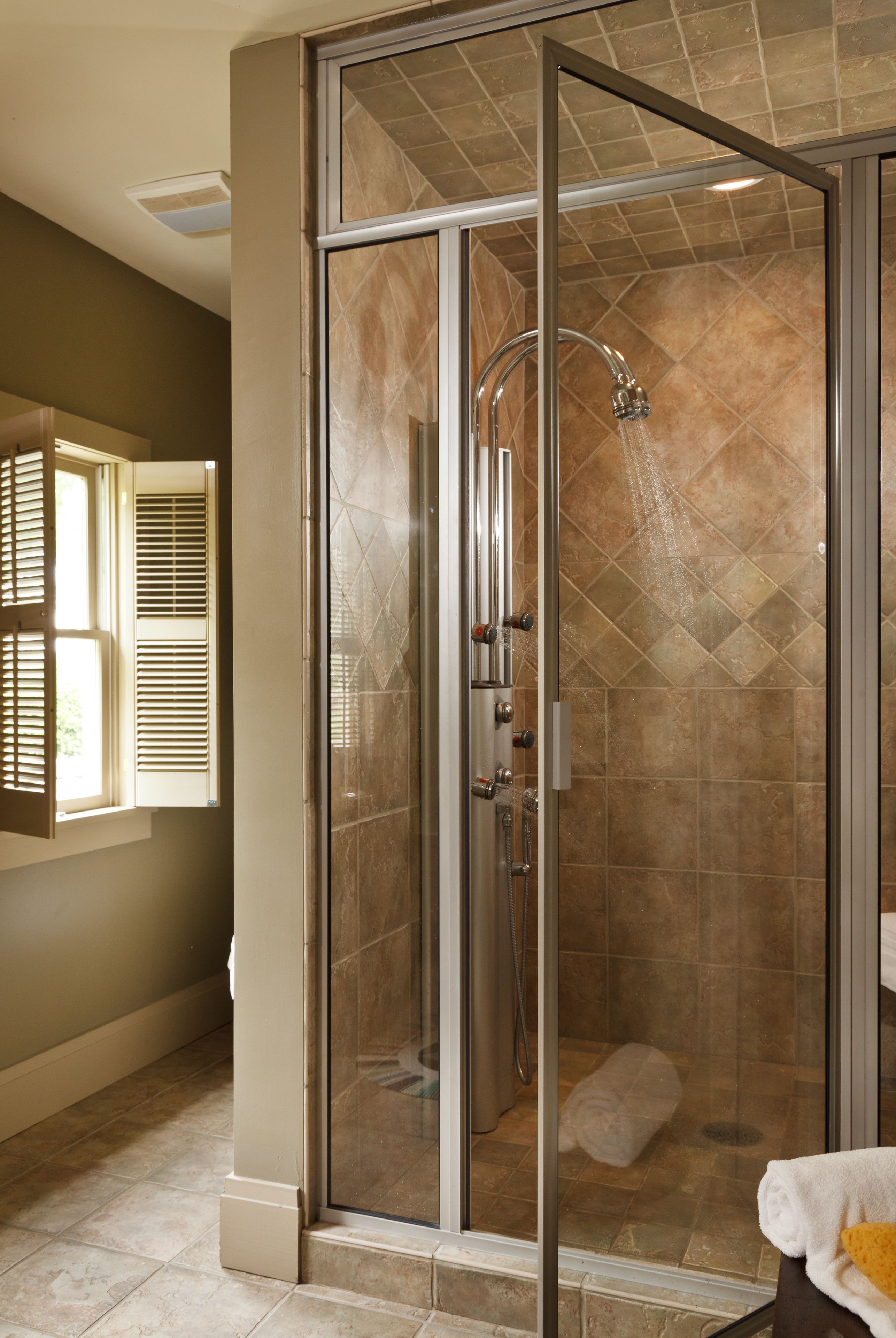 A glass-enclosed shower with a rain showerhead, a towel on the floor, and a window with blinds.