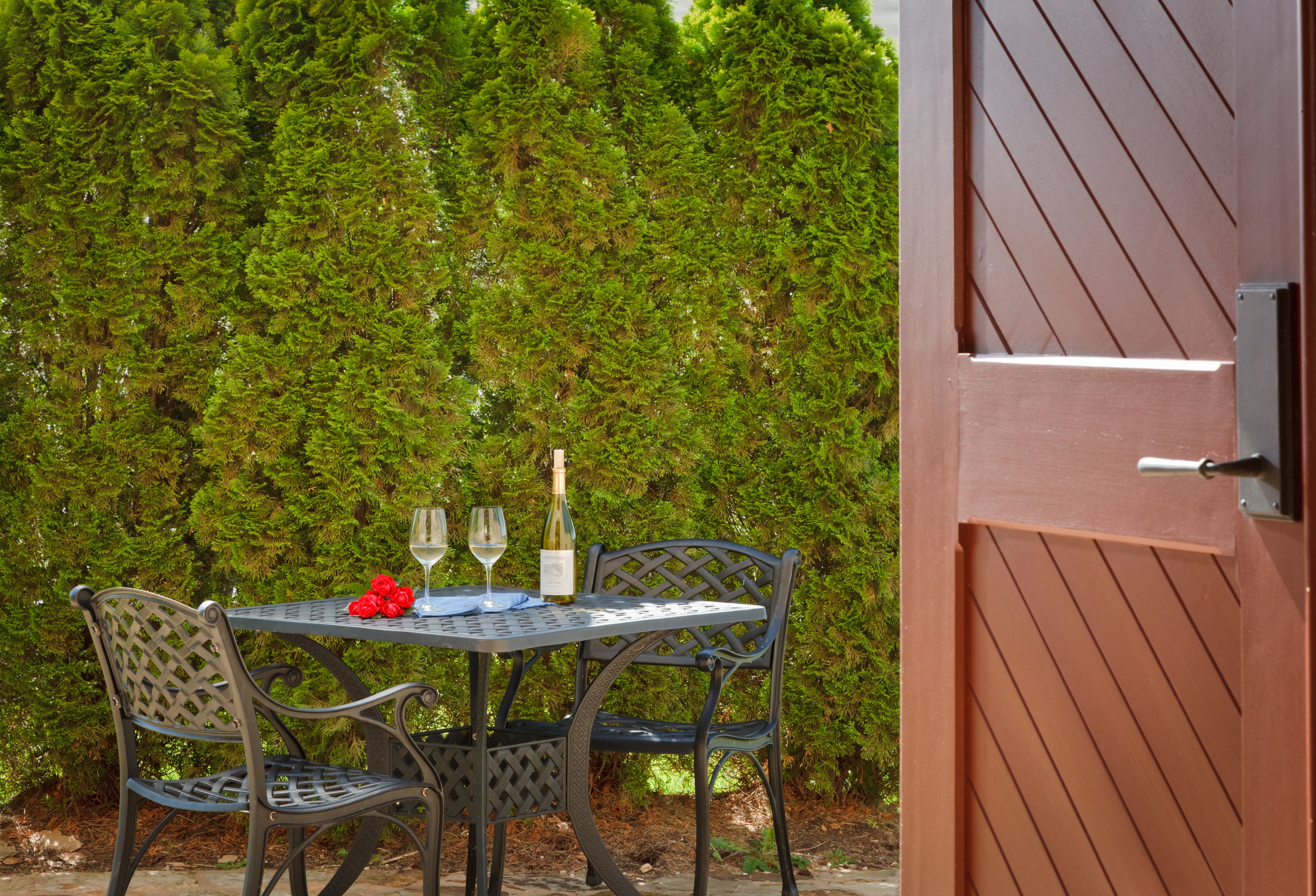Ein Tisch mit zwei Stühlen und einer Weinflasche, vor einer hohen, dichten Hecke.