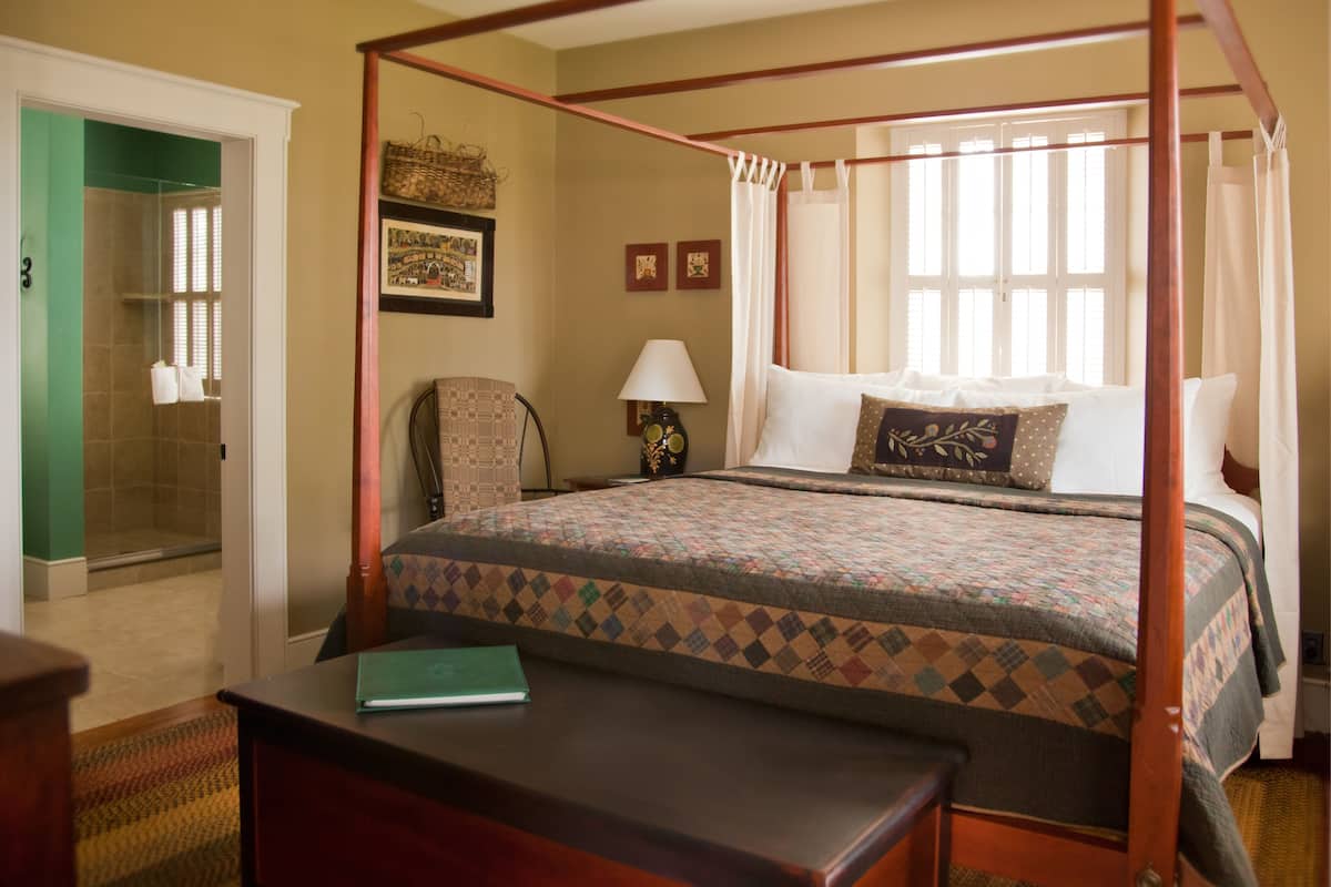A four-poster canopy bed with a quilt, a bedside table, a chair, a lamp, and a window with curtains.