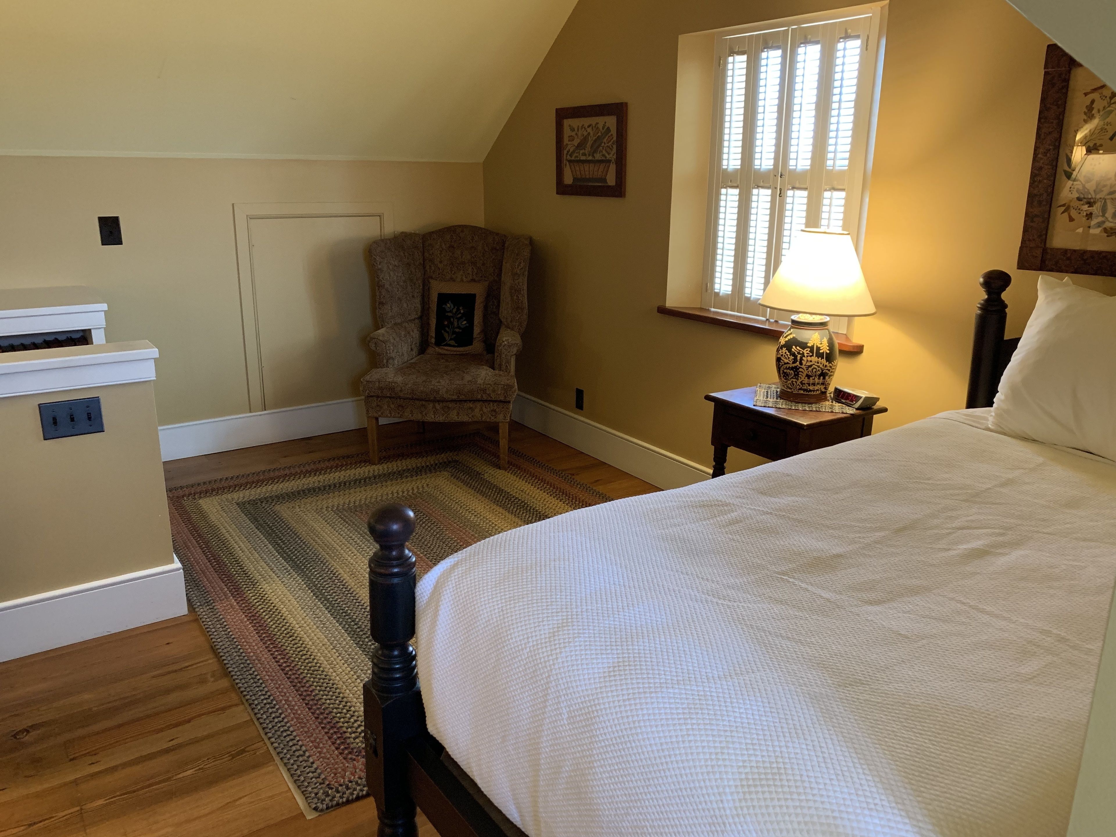 A bedroom with a bed, a chair, a nightstand, a lamp, and a window with blinds.