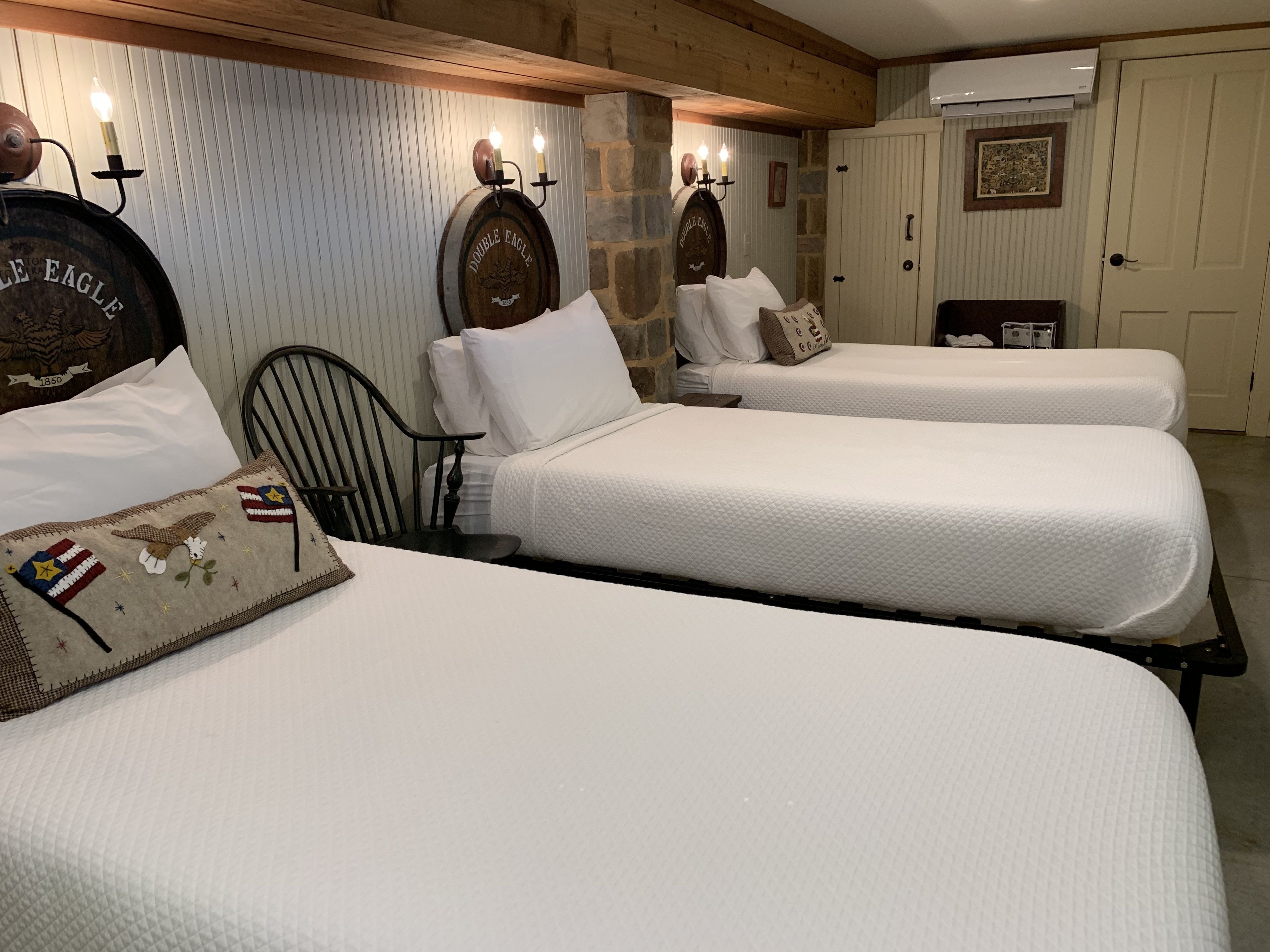 A room with three single beds, each with white bedding and decorative pillows. There are wall-mounted sconces with candles, a wooden headboard, and a stone accent wall.