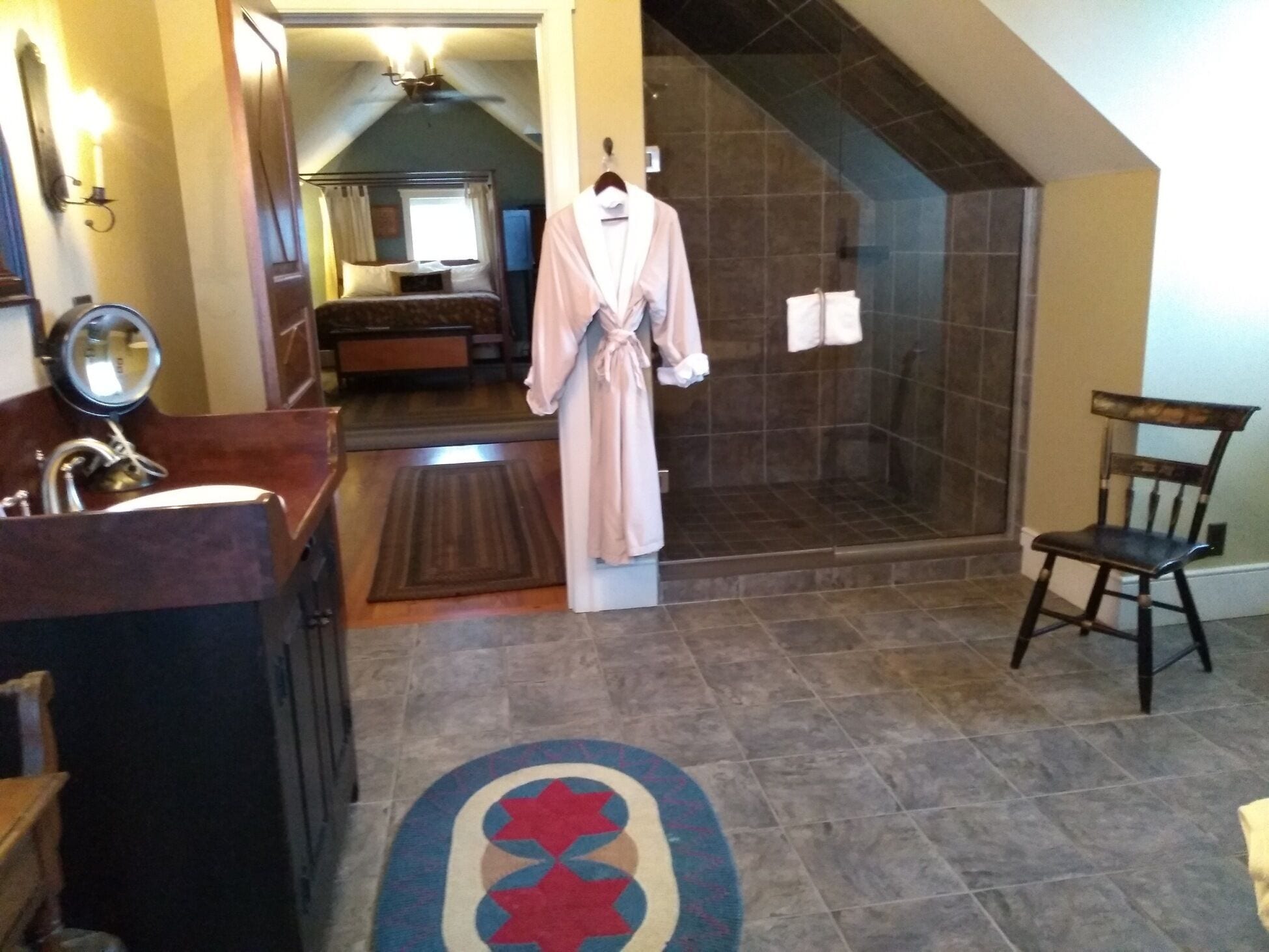 Un baño con ducha, una silla de madera, una bata colgada de un perchero y una alfombra estampada.