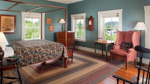 A bedroom with a four-poster bed, a red armchair, a wooden dresser, and a view of greenery through the windows. - Inn at Hermannhof (Hermann)