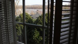 A view through a window with shutters, revealing a landscape with trees, a building with a tower, and a body of water in the distance. - Inn at Hermannhof (Hermann)