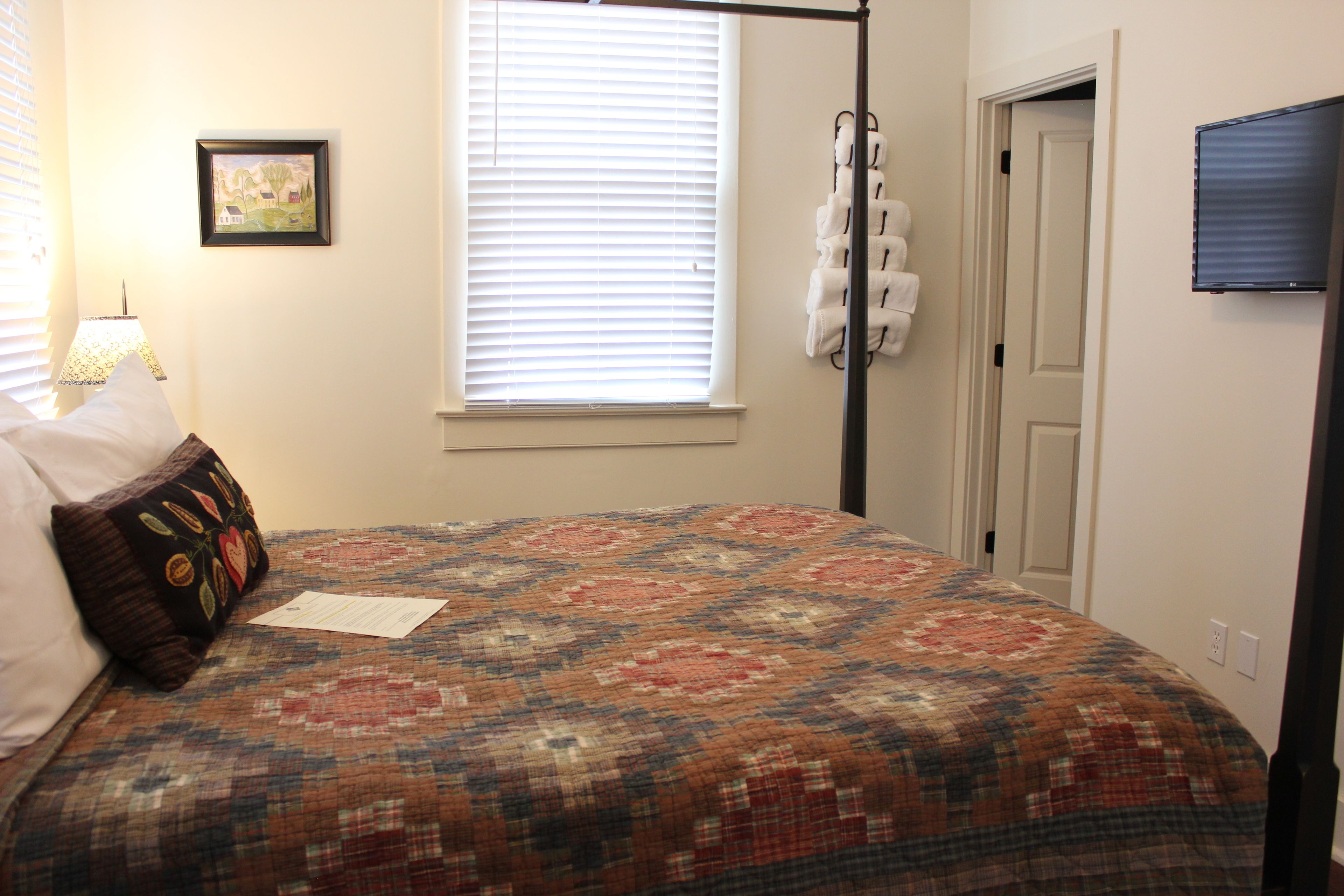 A neatly made bed with a patterned bedspread, a pillow with a leaf design, a framed picture on the wall, and a window with blinds.