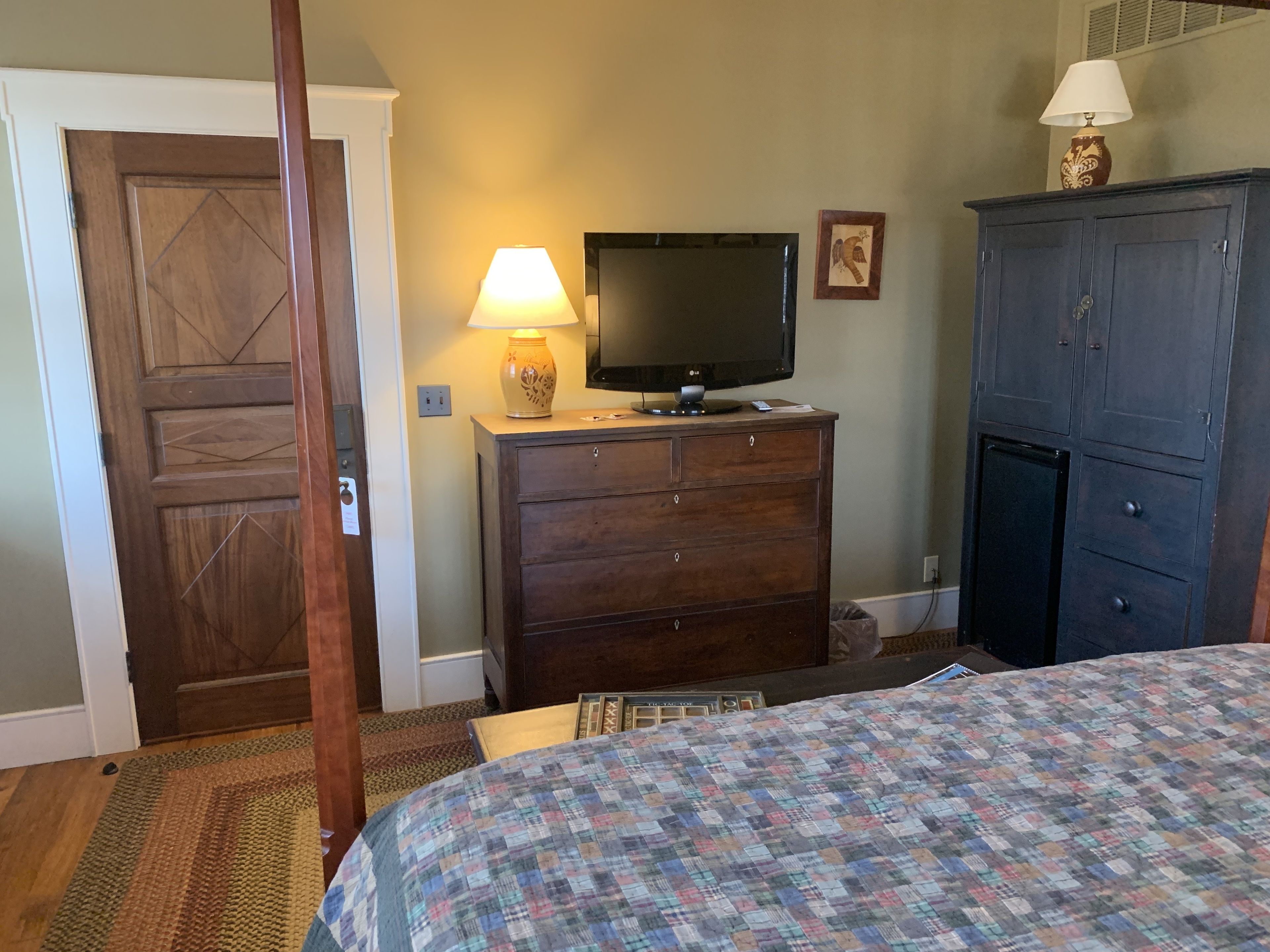 A bedroom with a wooden door, a dresser with a TV, a lamp, and a bed with a quilt.