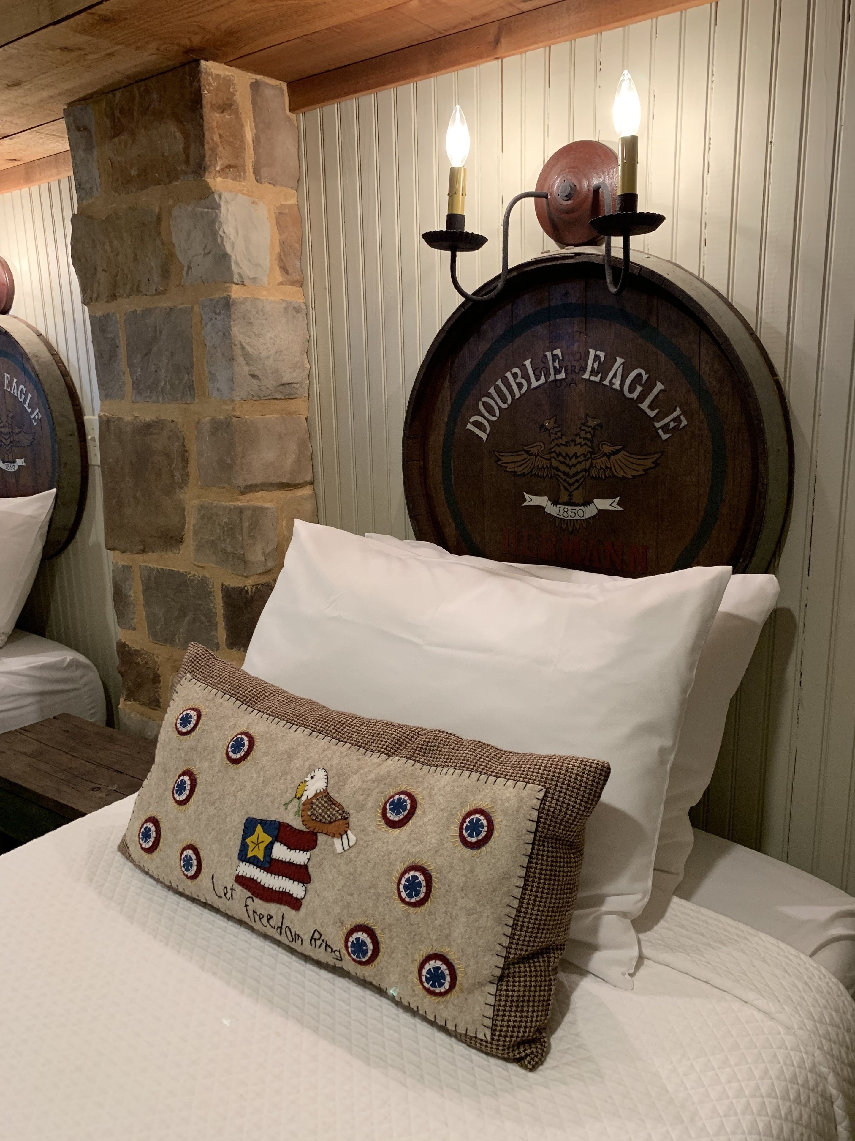 A bed with a quilted pillow featuring a patriotic design, a stone wall, and a wooden ceiling.