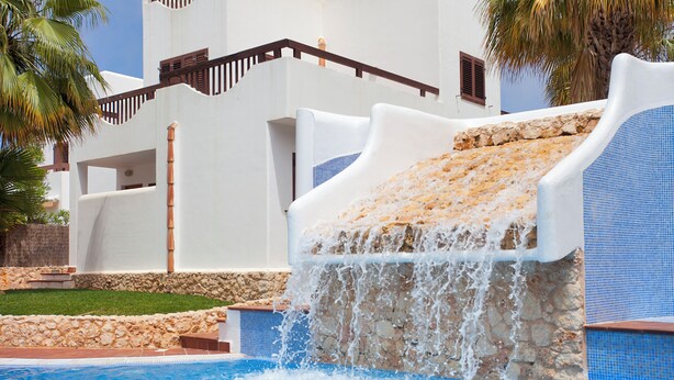 A pool with a waterfall feature, stone walls, and a white building with balconies. - Marina dór IV, house 5, wifi 5 bedrooms, community pool, beach 300m away (Santanyí)