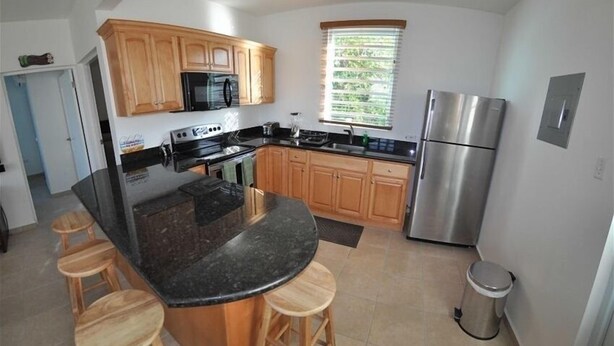 A modern kitchen with wooden cabinets, a stainless steel refrigerator, a black countertop island with stools, and a window with blinds. - High End Amenities, Panoramic Sunset View, Roof Top Deck (Aguada)