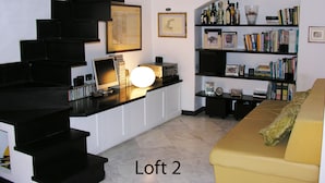 En hems med trappe, et skrivebord med computer, en reol og en gul sofa.