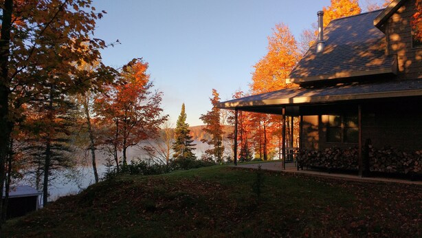 Un chalet au bord du lac, doté d’un patio couvert et offrant une vue sur le feuillage automnal et le lac.