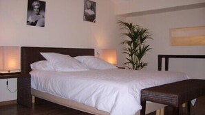 Una cama bien hecha con sábanas blancas, cabecera de madera y mesita de noche con lámpara. Hay dos cuadros enmarcados en la pared sobre la cama.