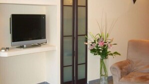 Una sala moderna con un televisor de pantalla plana sobre un estante blanco, una puerta de vidrio, un jarrón con flores y un sillón color beige.