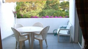Un patio techado al aire libre con una mesa redonda y cuatro sillas, un pequeño sofá blanco, y vistas a follaje y flores.