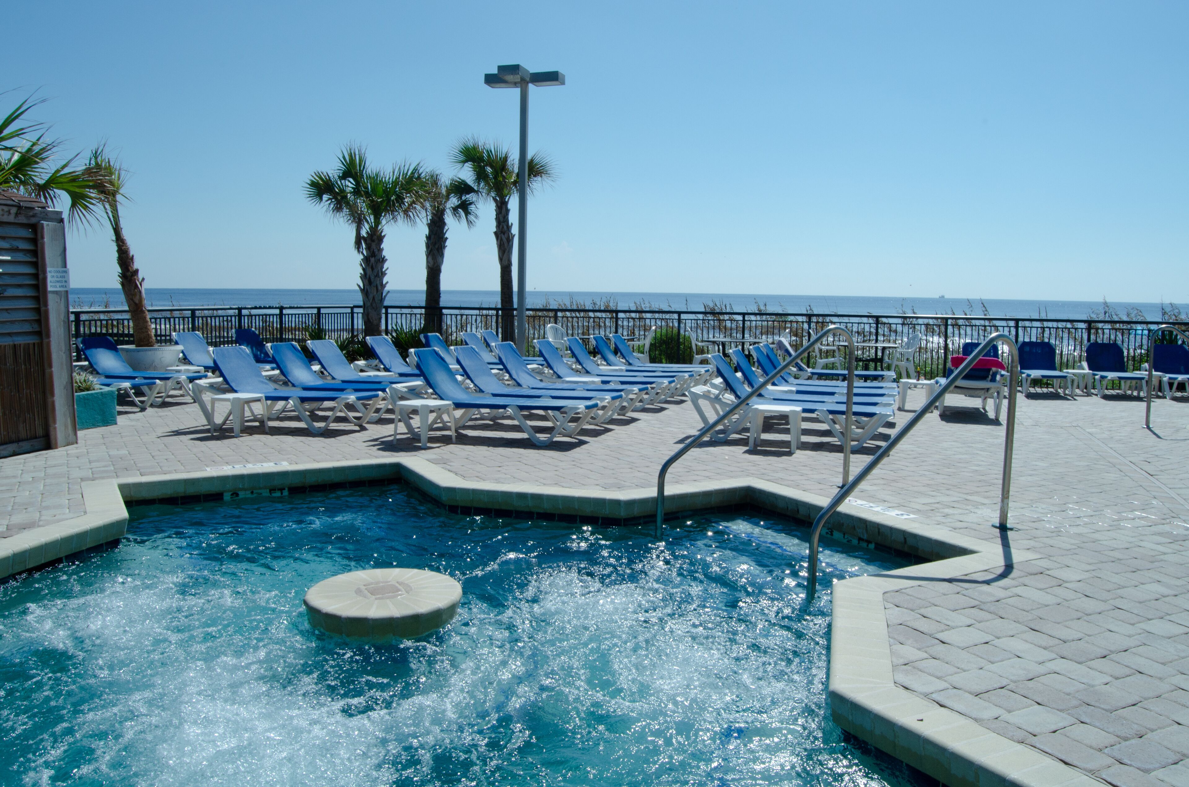 A pool area with blue lounge chairs, a hot tub, and a view of the ocean.