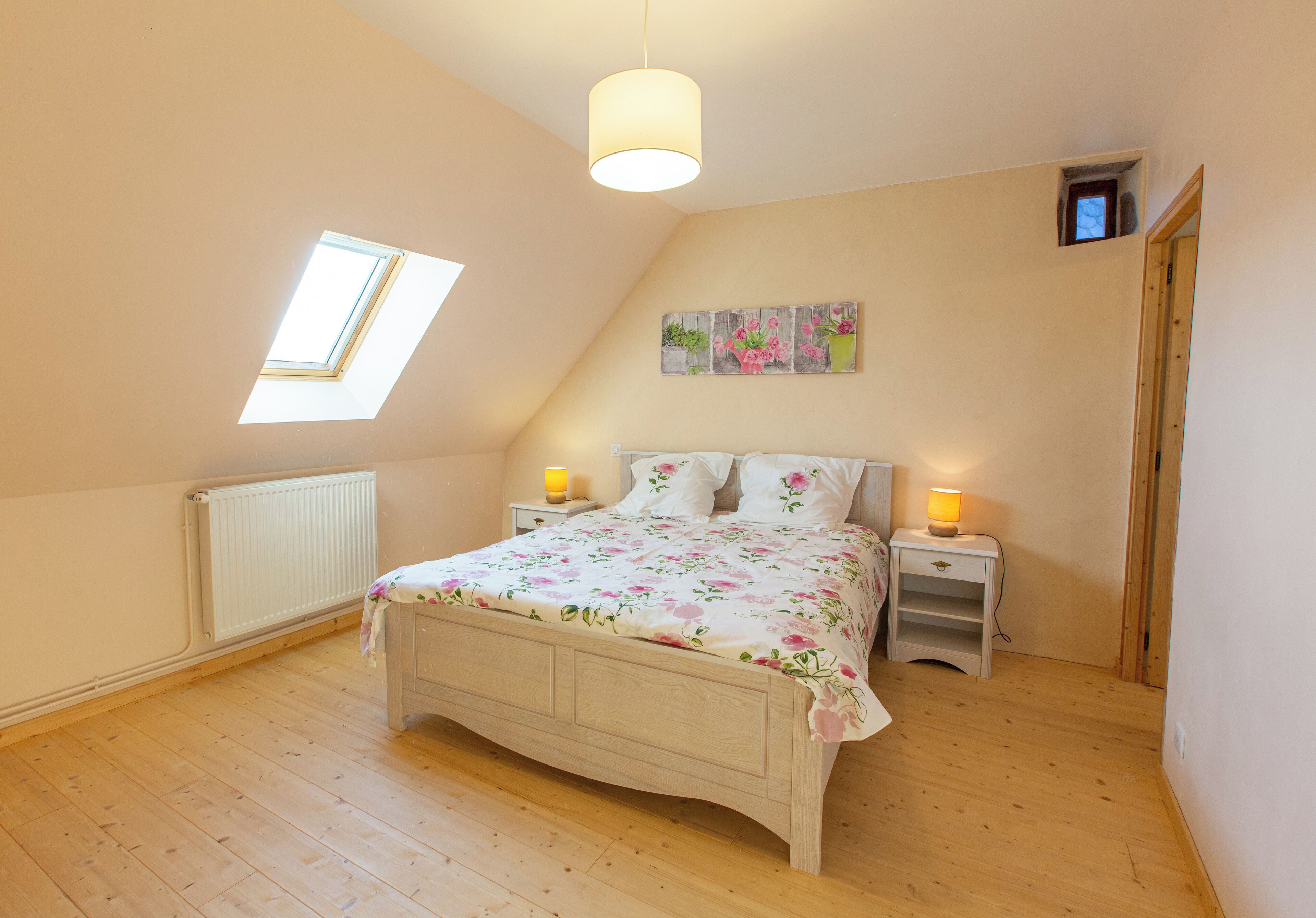 Quarto com teto inclinado, claraboia, cama com roupa de cama florida, luminárias de cabeceira e um pequeno quadro na parede.