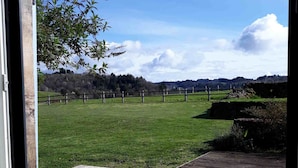 Blick aus einem Fenster auf eine Holzterrasse und ein grünes Feld mit einer Steinmauer und einem Zaun.