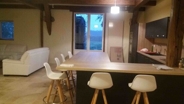 A modern kitchen with a wooden countertop and bar stools, a white sofa, and a large window with a view. - Typical Corrèze barn in stone in a preserved nature (correze)